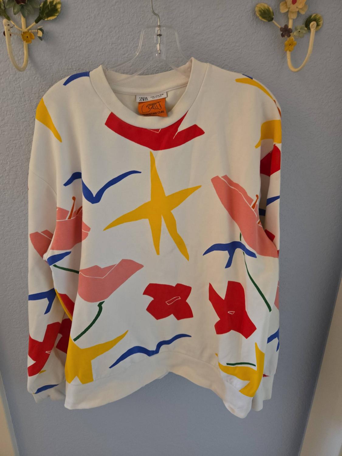 White sweatshirt hanging with an abstract print of stars and colourful shapes. ZARA and SEBASTIAN CURI labels at the neck.