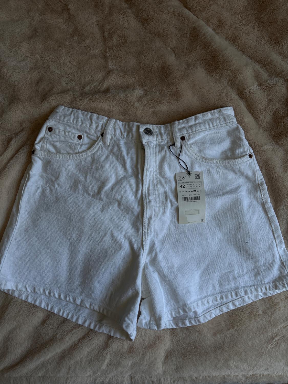 White denim shorts with a size 42 label, laid out on a soft beige blanket. Casual and relaxed style.