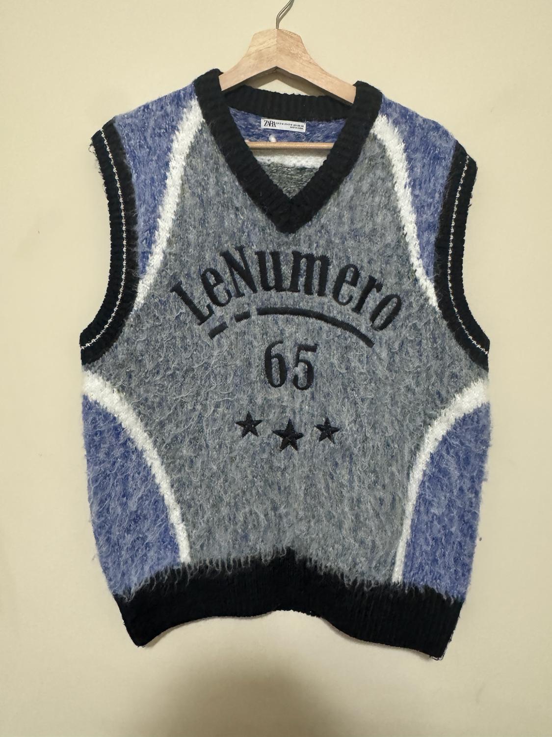 Gray and blue knit vest on a hanger. Black V-neck, ZARA label, text "LeNumero 65" and three stars. Light background.