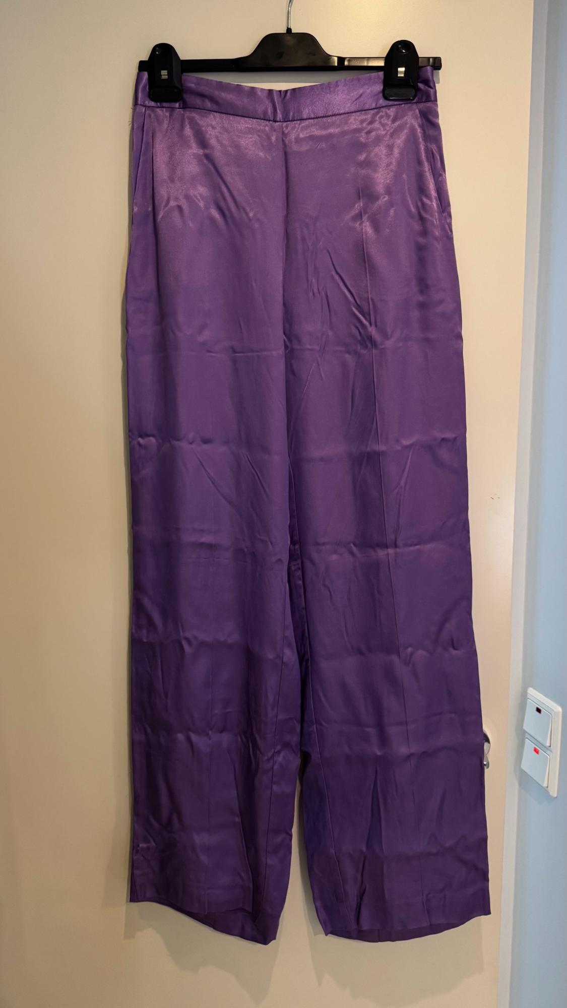 Purple wide-leg trousers in a shiny fabric hanging on a black hanger over a light-coloured door. To the right is a light switch.