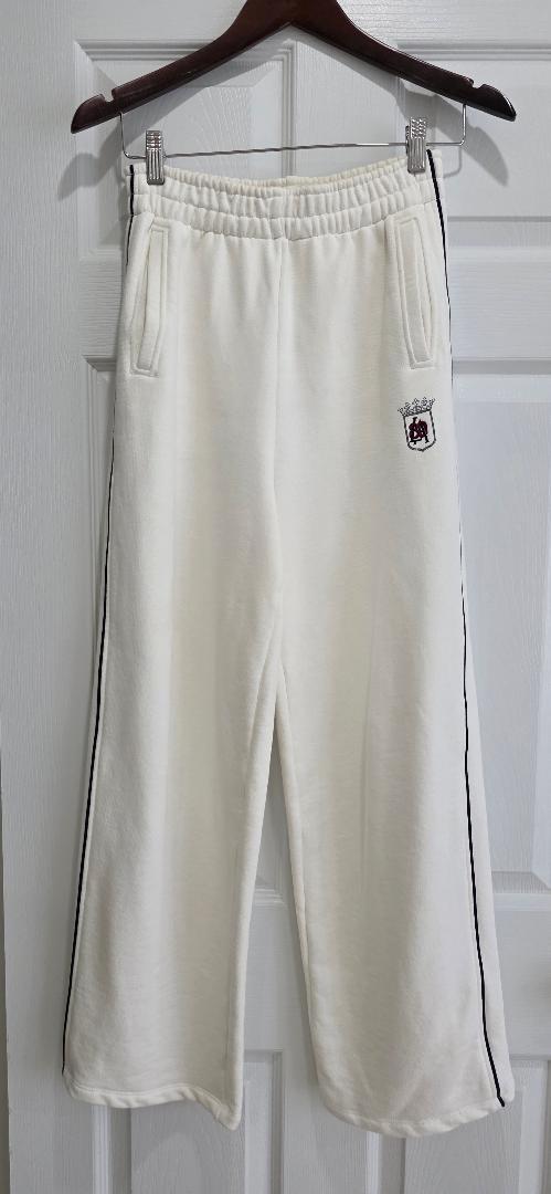 White fleece pants hanging on a hanger, with black stripes on the sides and an embroidered emblem on the top left.