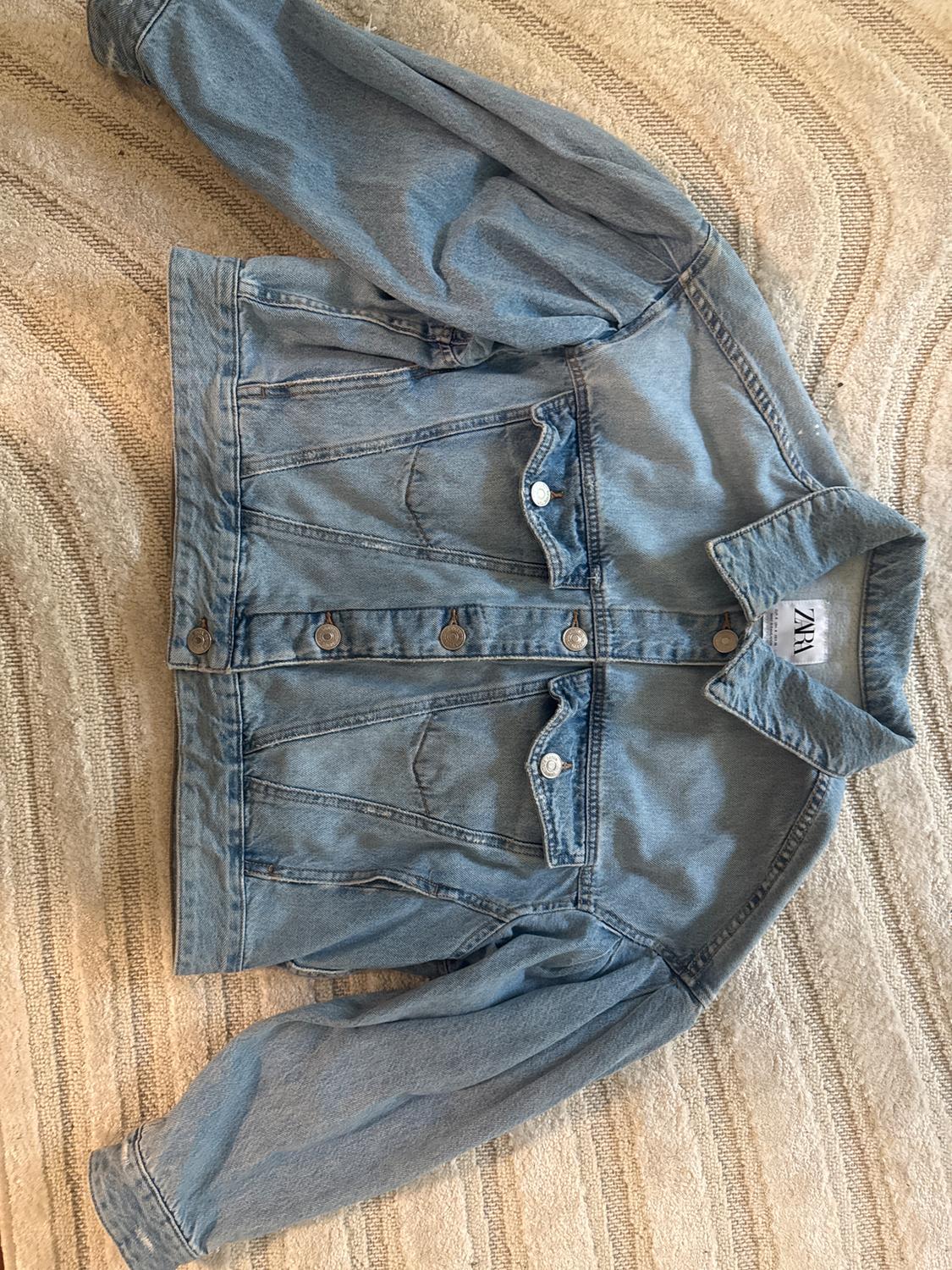 Light blue denim jacket with metallic buttons and front pockets, on a beige rug with a curved line pattern. Visible tag: ZARA.