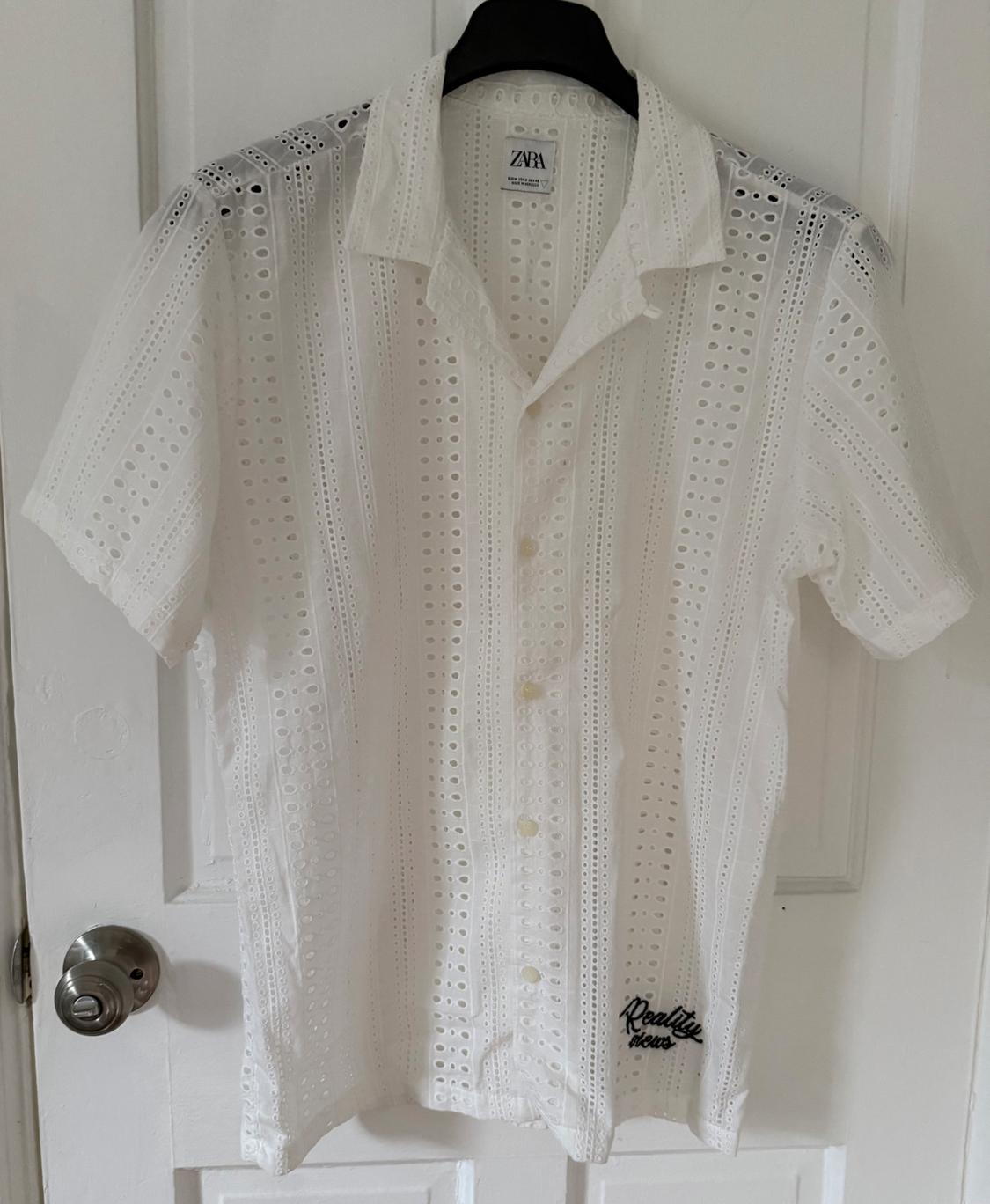 White perforated short-sleeved shirt hanging on a door. ZARA label and the text Reality news on the hem.