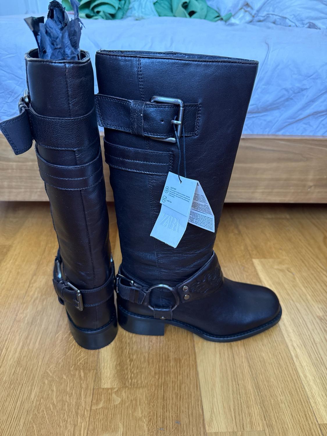 Black high boots with straps, buckles and ZARA and LEATHER labels on a wooden floor next to a bed.