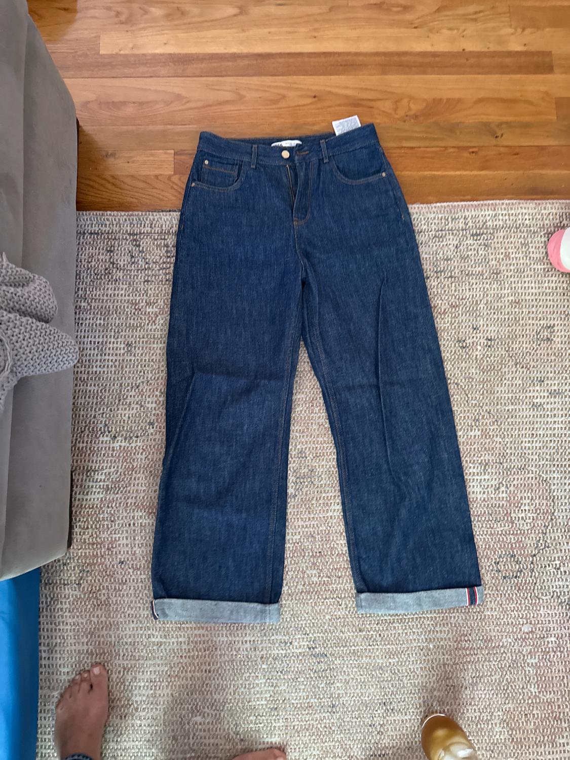 Dark blue jeans with a hem, placed on a beige rug. Part of a sofa and feet visible in the image.