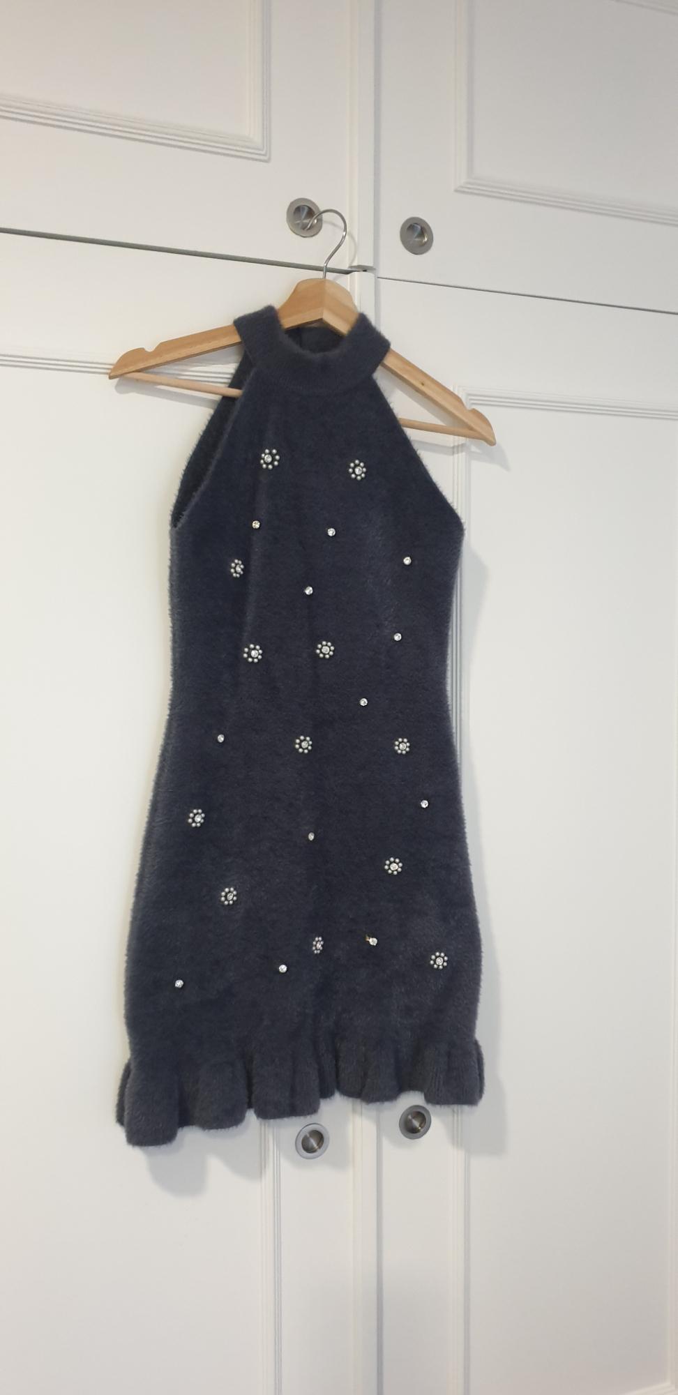 Sleeveless black dress with a high neck, decorated with pearls and flowers, hanging on a wooden hanger in front of a white wardrobe.