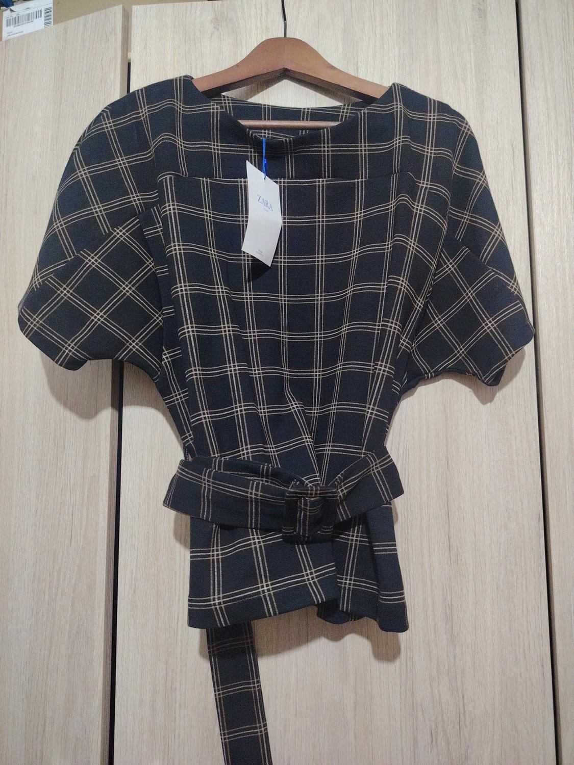 Dark checked blouse with a matching belt, hanging on a wooden hanger. Visible label with the text "ZARA". Light wooden background.