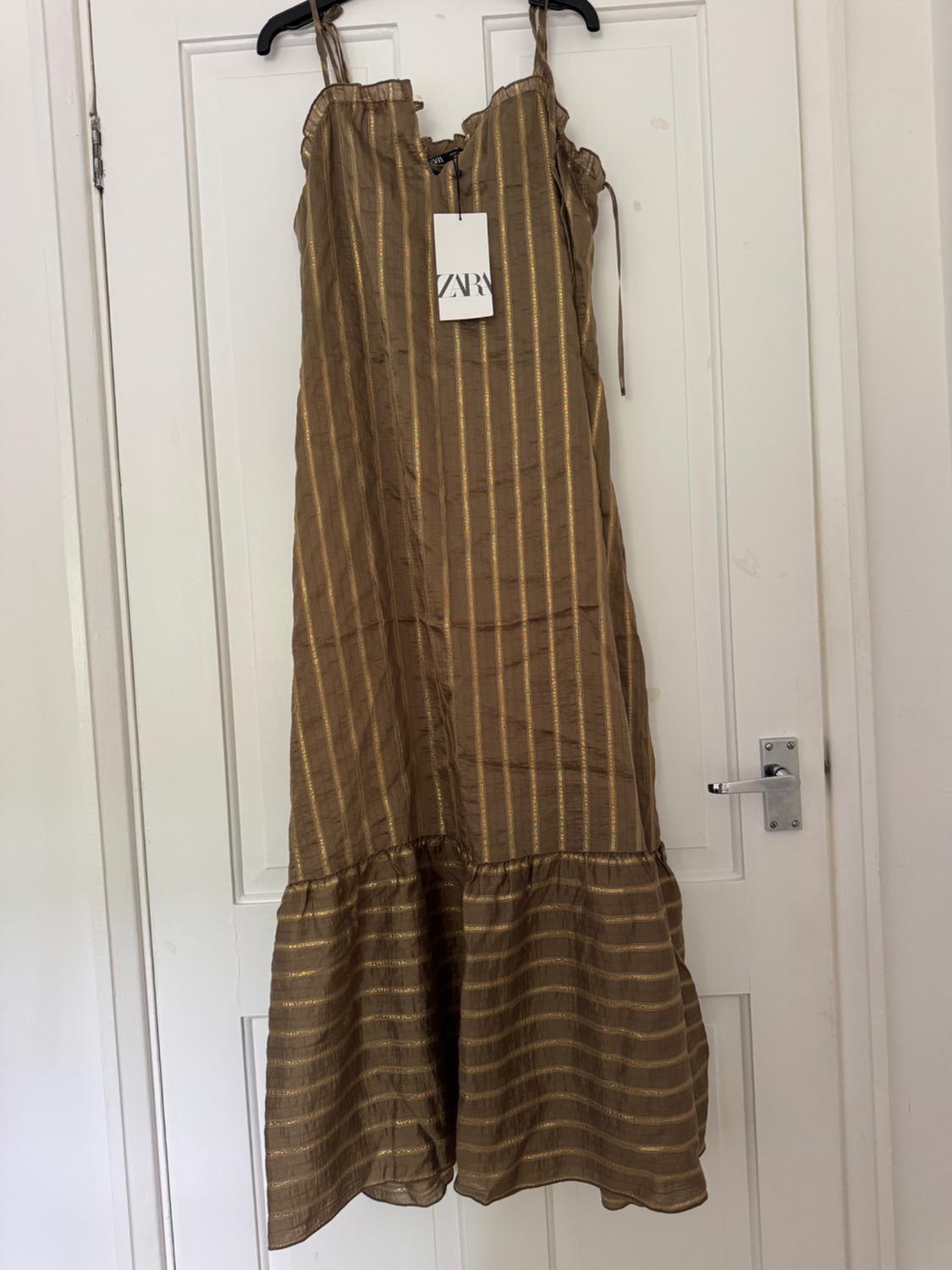 Brown strappy maxi dress with gold stripes and a frill, hanging on a white door. Label with the text ZARA.