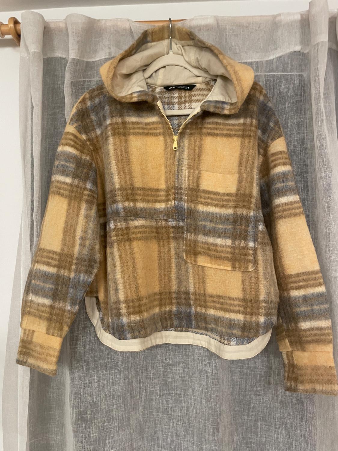 Brown and beige plaid hooded jacket, hanging on a hanger over white and gray curtains.