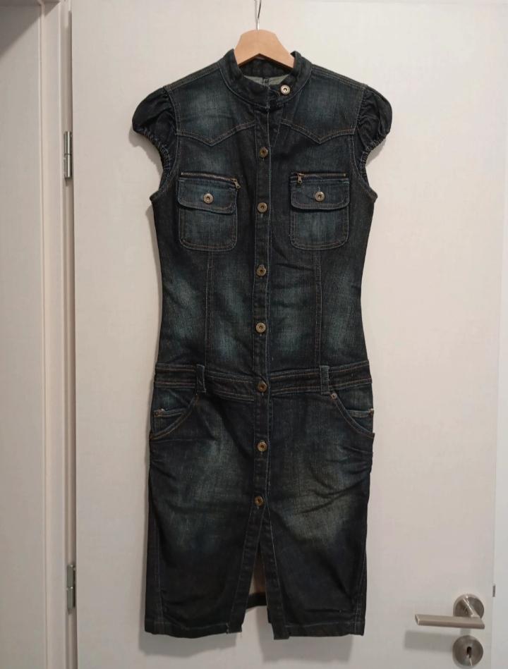 Sleeveless denim dress hanging on a wooden hanger. It has front buttons and chest and hip pockets.