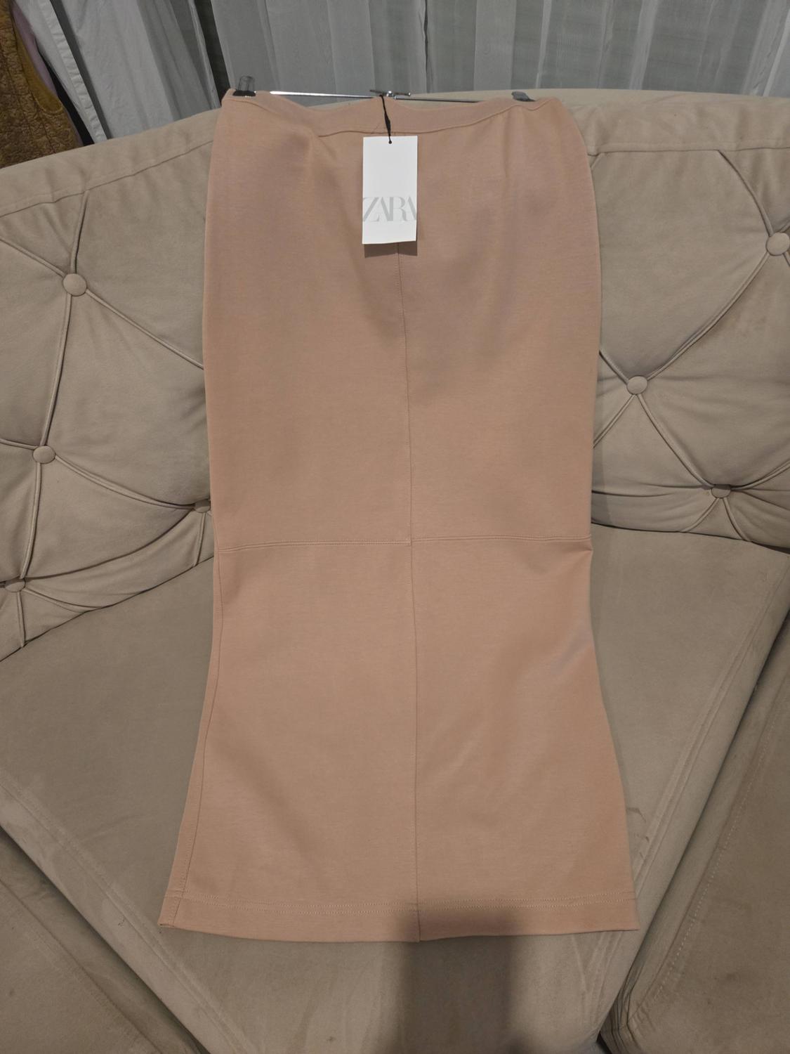 Long beige skirt with stitching, laid out on a buttoned sofa. There is a white label on the waistband with the text ZARA.