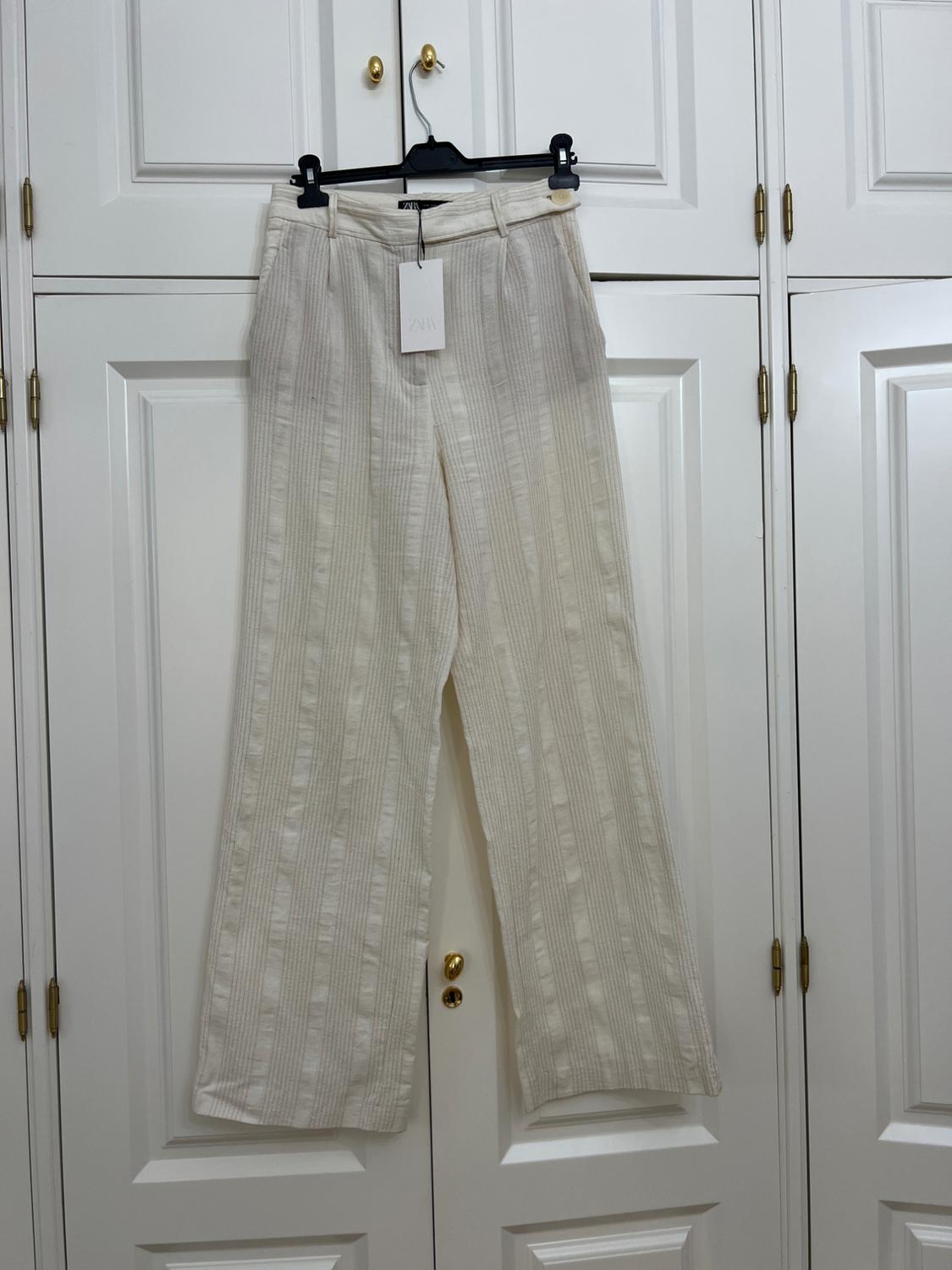 Light-coloured vertical striped trousers hanging on a black hanger over the doors of a white wardrobe. Label with the text ZARA.