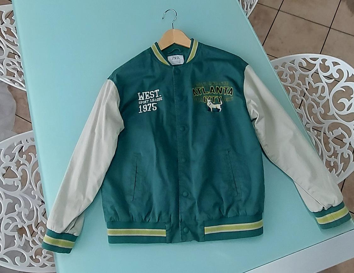 Green jacket with white sleeves on a blue table. Text: "WEST SPORT LEAGUE 1975" and "ATLANTA CITY". Decorative white chairs around.