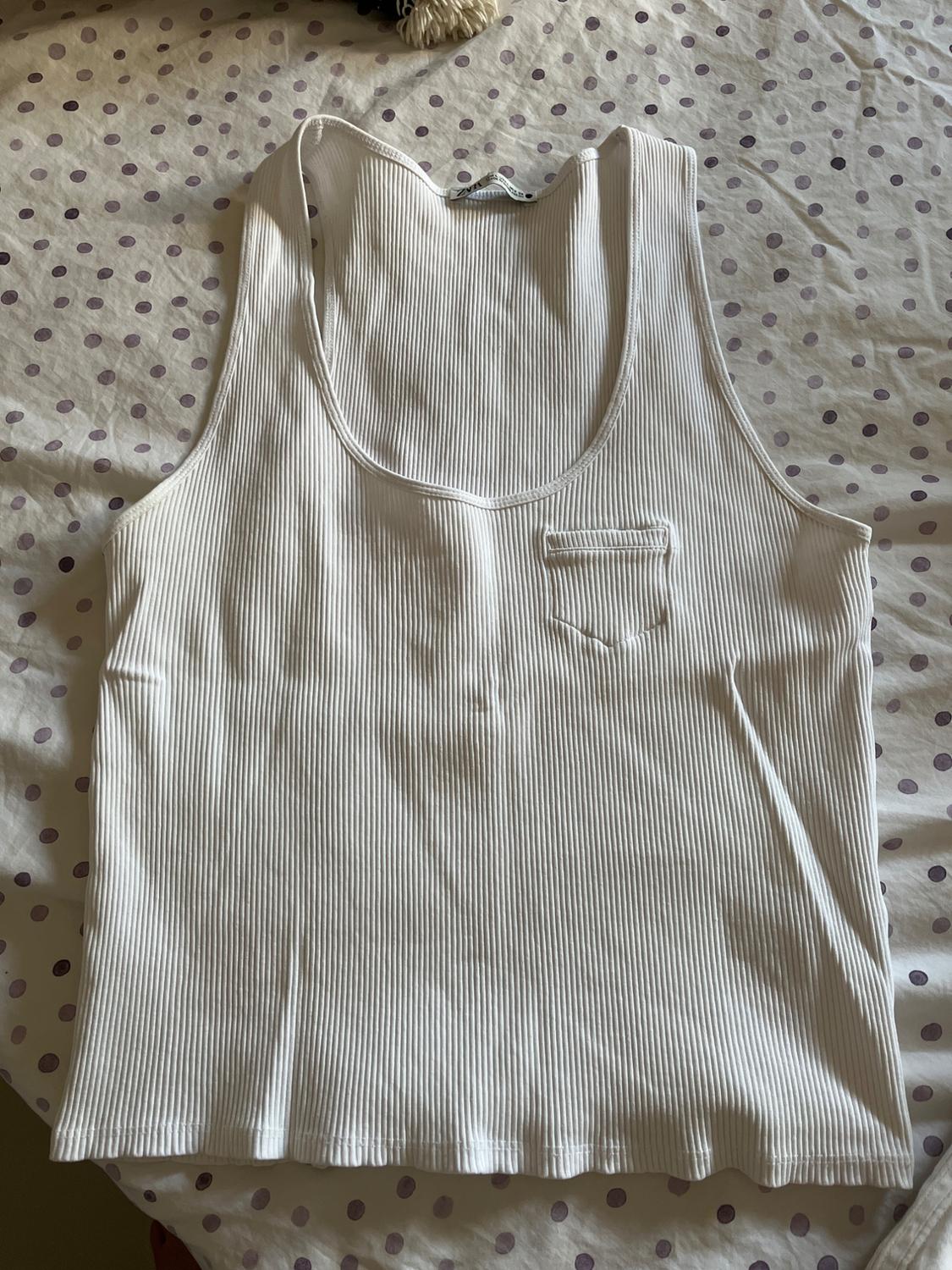 White ribbed vest top with a small chest pocket, set against a polka dot fabric background. ZARA label at the neck.