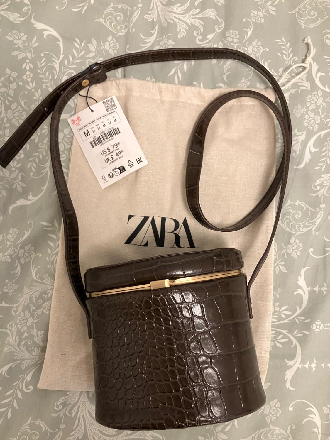 Brown crocodile-texture bag on a fabric shopping bag with Zara text and a label reading M US $ 79.90 UK £ 49.99. Floral background.