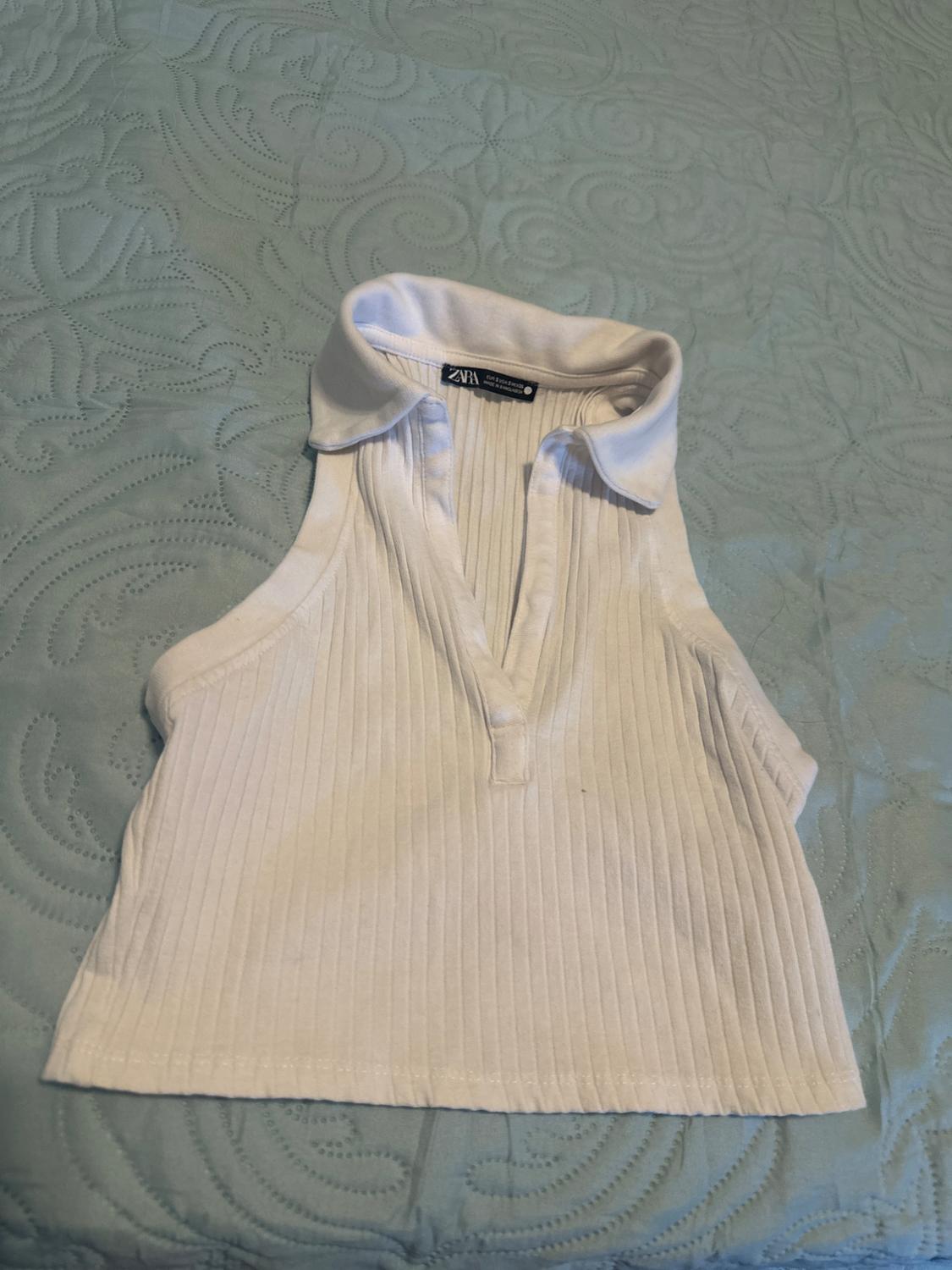 White ribbed sleeveless cropped top with a polo neck and V-neckline, featuring a ZARA label, laid on a textured light blue bedspread.