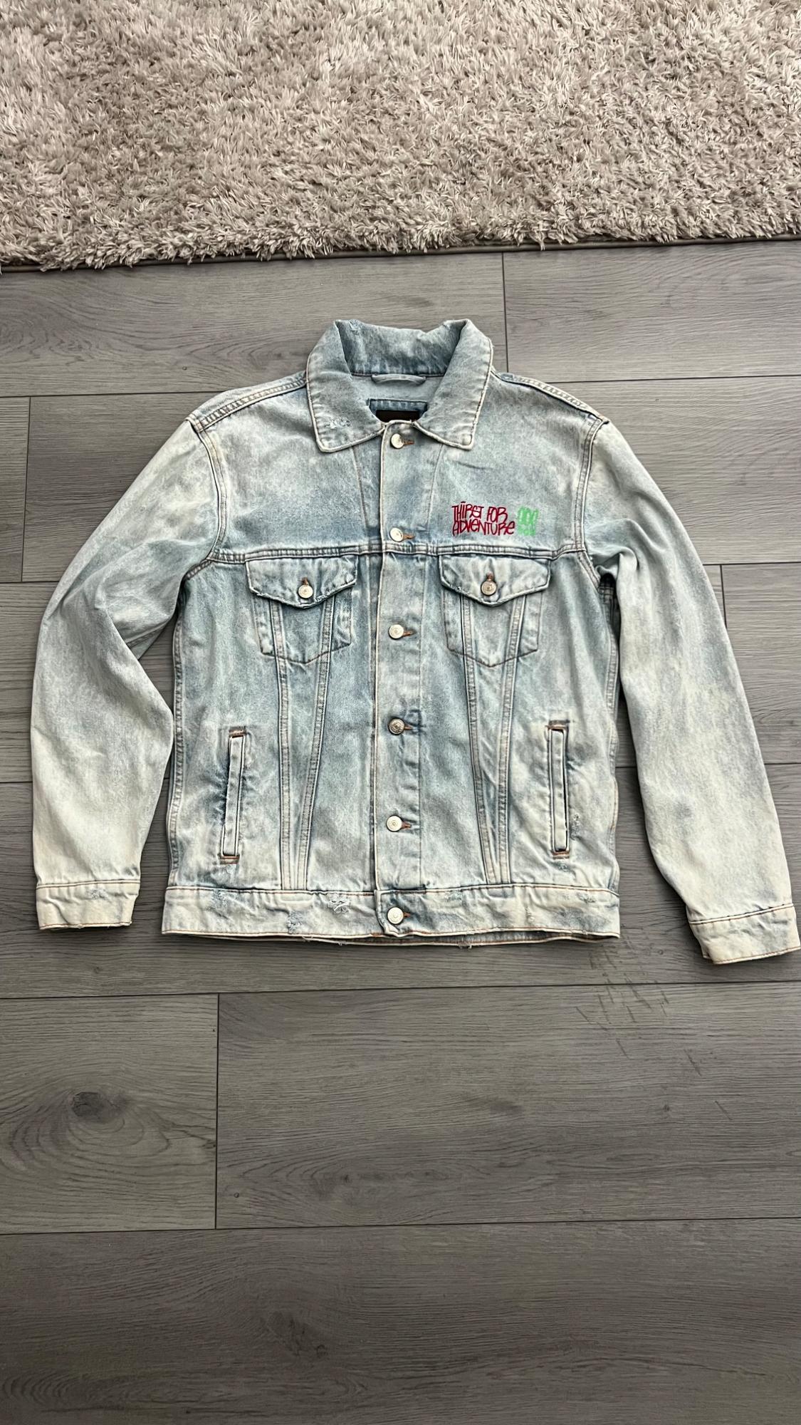 Light blue denim jacket on a gray wooden floor. On the left chest, it has the text THIRST FOR ADVENTURE. Casual style.