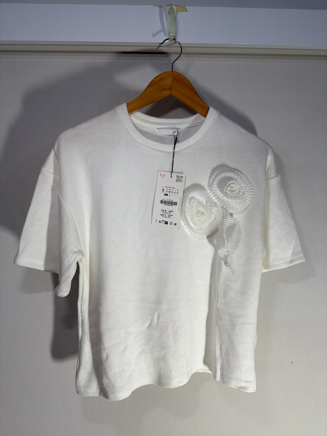 White short-sleeved t-shirt with embossed floral detail, hanging on a wooden hanger. Visible tag with price and size.