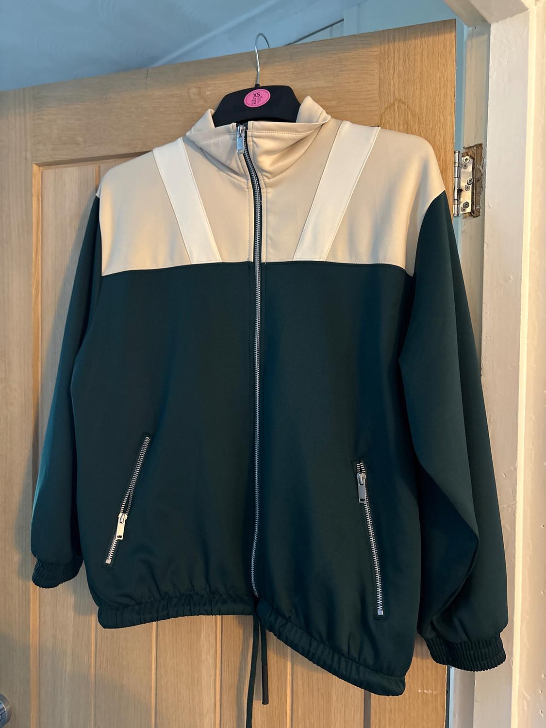 Jacket hanging on a door, with a front zip. Beige top and dark green bottom, with side pockets.