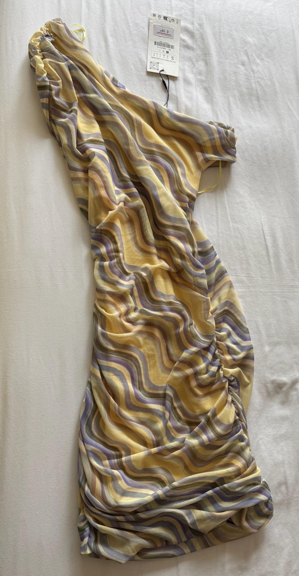 Asymmetrical dress with a wavy print in yellow and lilac tones, featuring side ruching and a label, on a white surface.