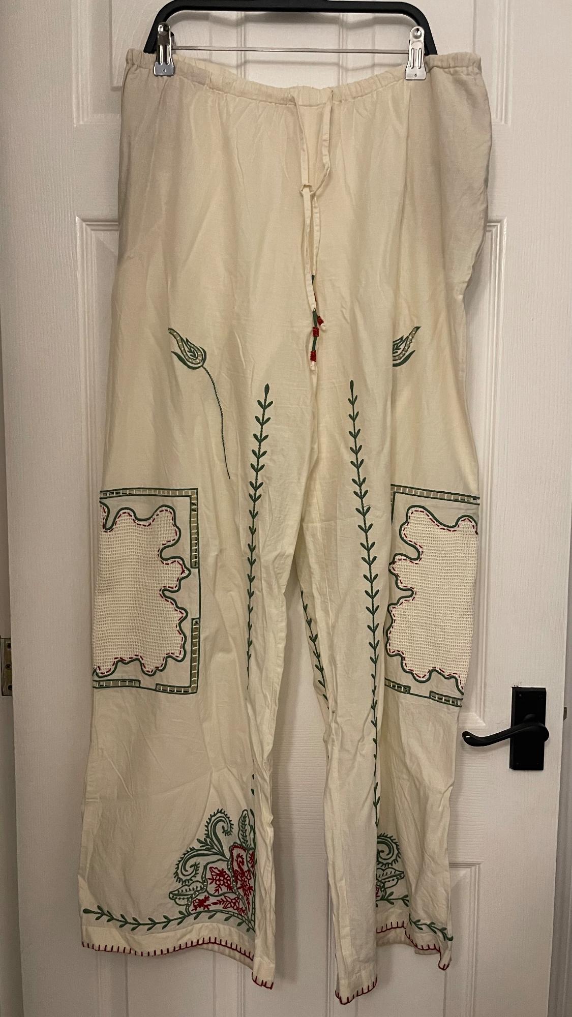 White trousers hanging on a door, with green and red leaf and flower embroidery, and decorative side pockets.
