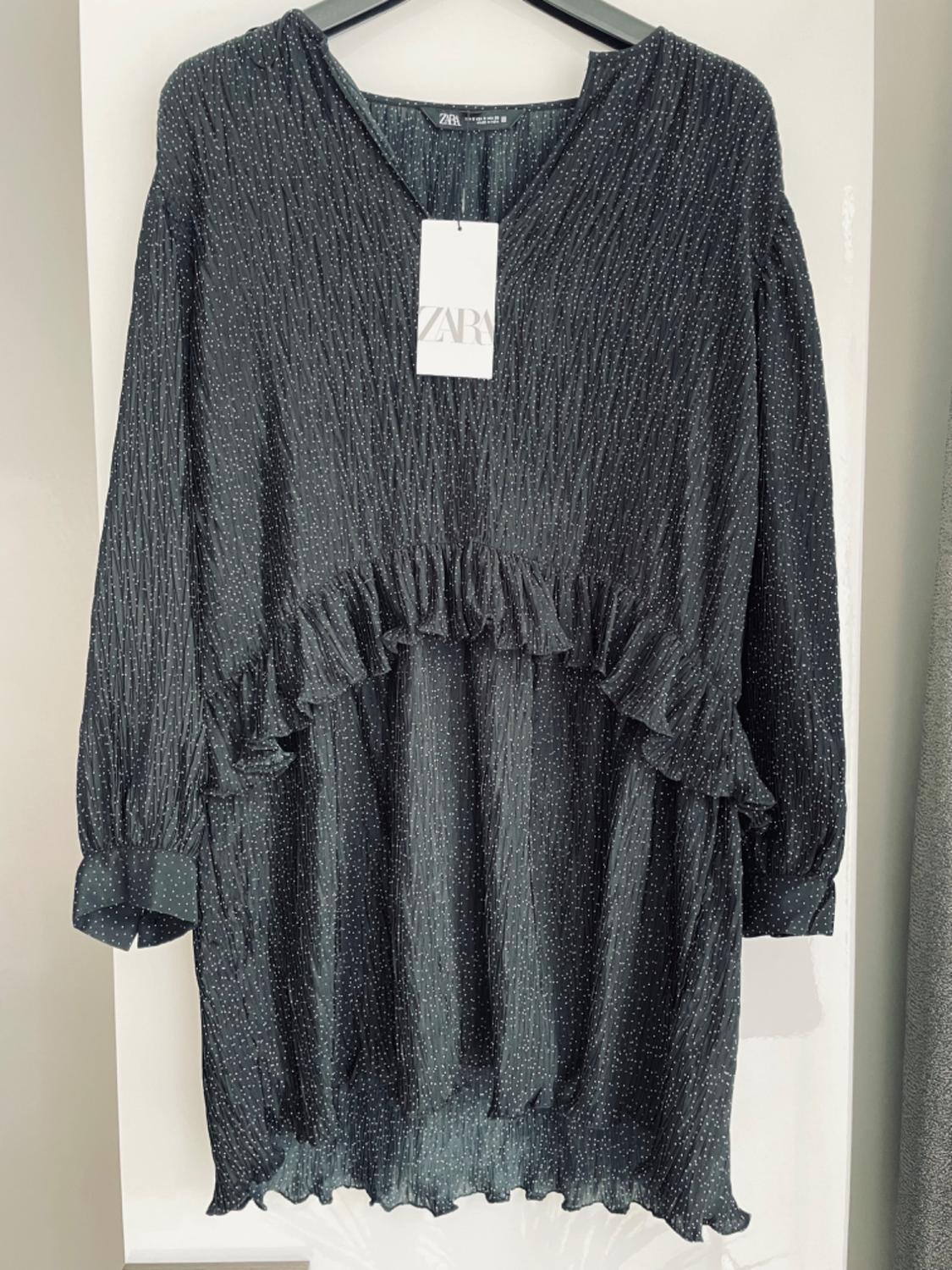 Front view of a black dress with frills and white dots, V-neck, long sleeves and visible tag.