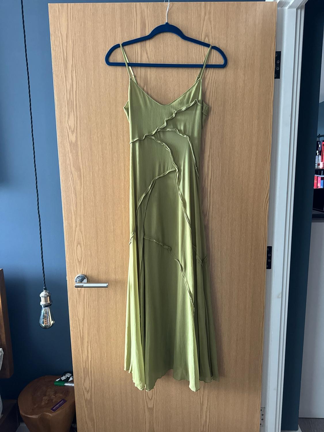 Green strappy dress with asymmetrical seams hanging on a wooden door. Blue wall and stool with the text APERITIVO.
