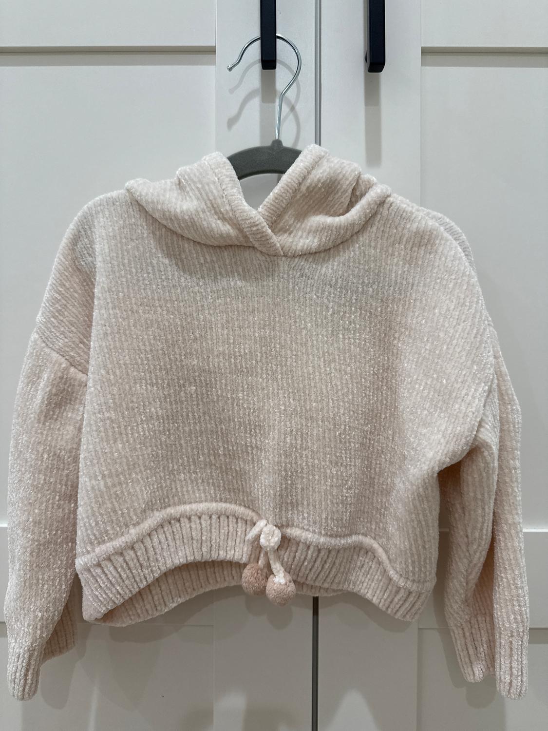 Beige knitted jumper with a hood and pom-poms on the hem, hanging on a hanger in front of a white door.