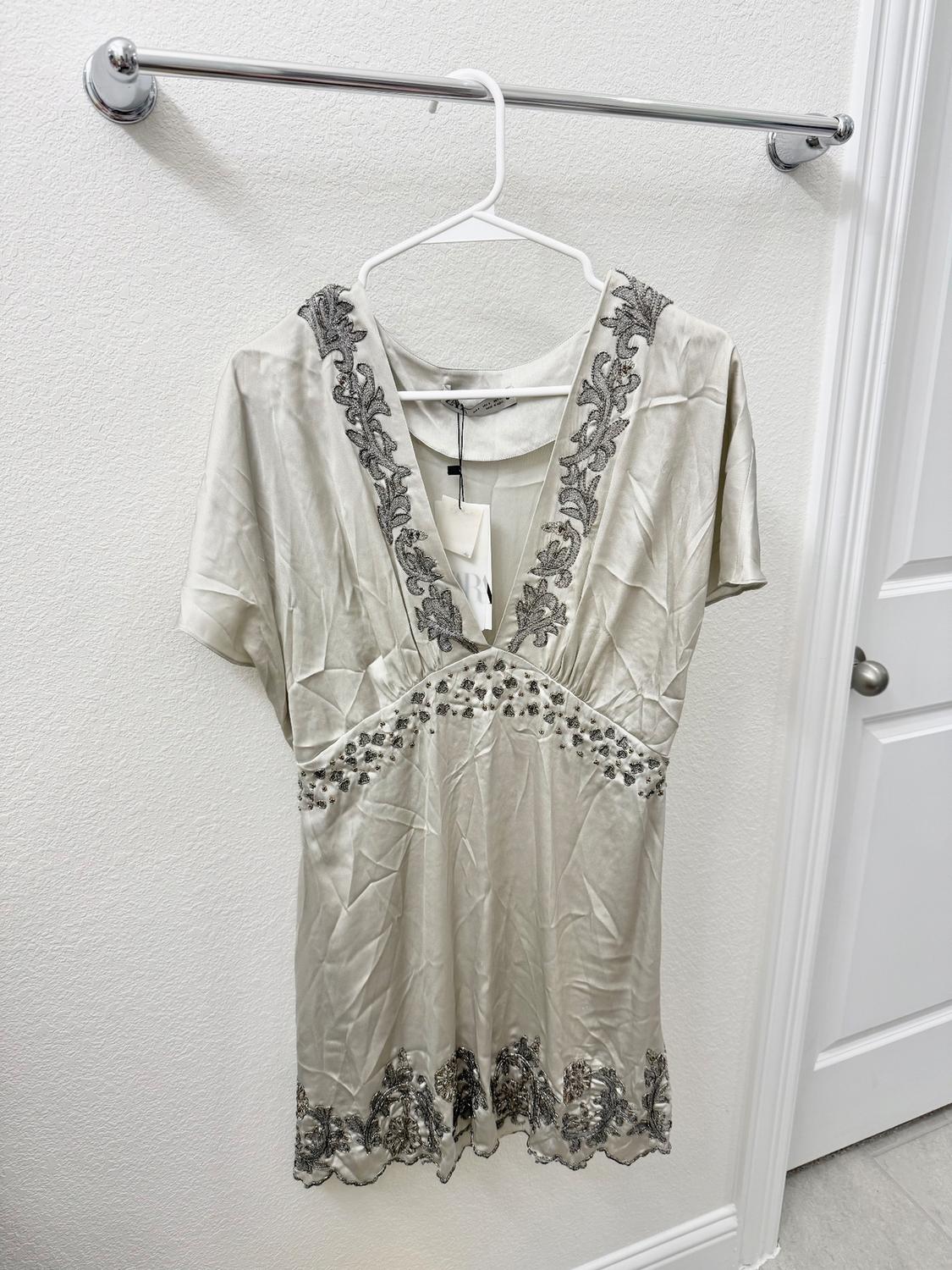 Beige dress with black floral embroidery on the neckline and hem, hanging on a white hanger against a white wall.