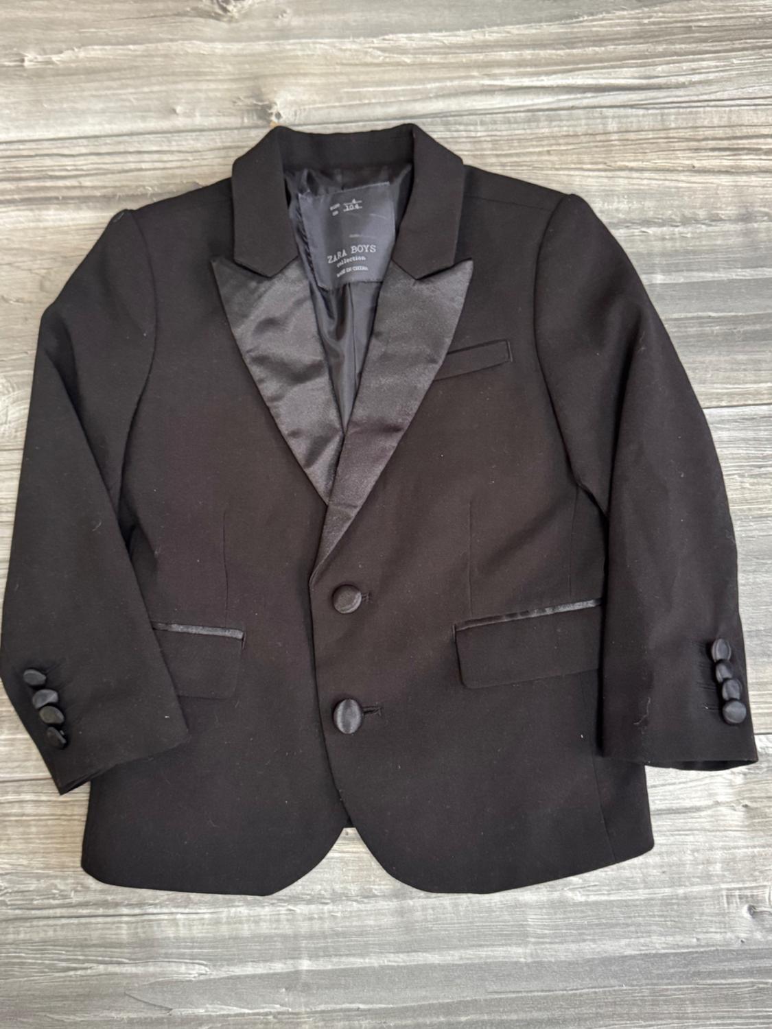 Black jacket with satin lapels against a wooden background. Label with the text ZARA BOYS collection, size 4 cm 104.