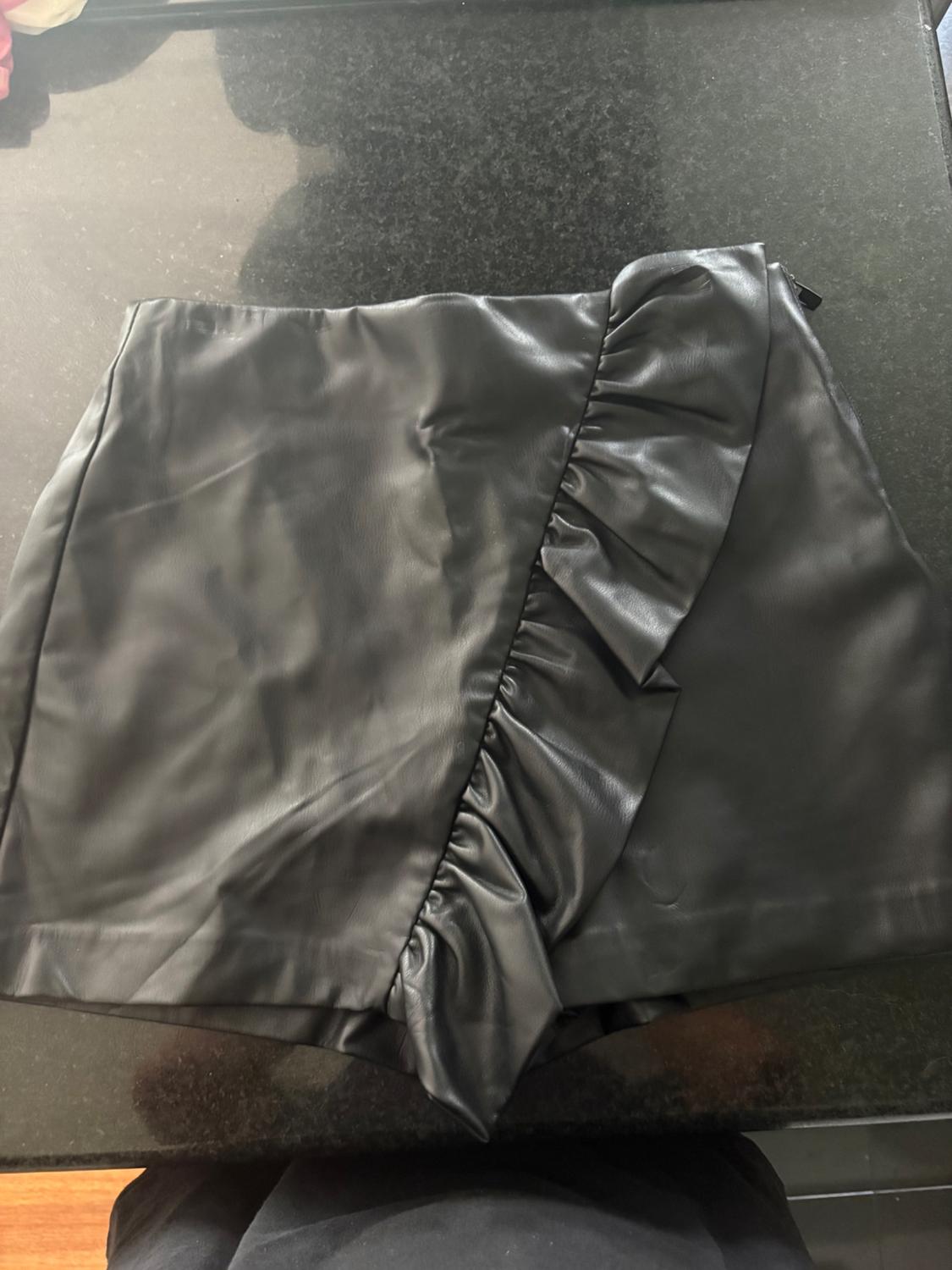Black skort with a glossy finish and an asymmetrical front ruffle, laid on a dark surface.