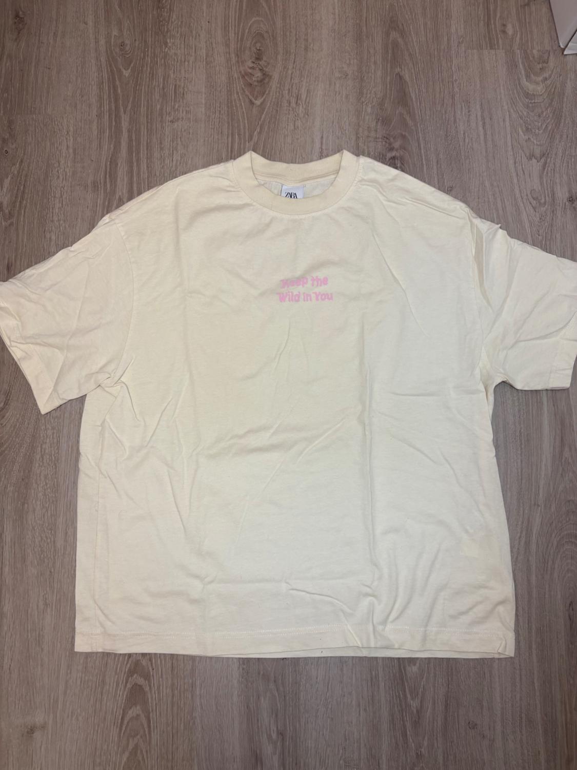 Beige T-shirt on a wooden floor, with pink text on the chest that reads "Keep the Wild in You".