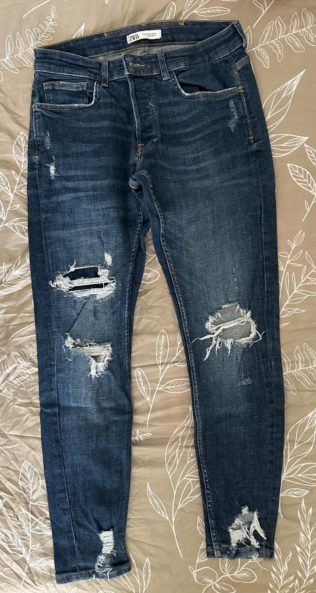 Blue jeans with ripped details and ZARA label, on a beige background with a white leaf print.