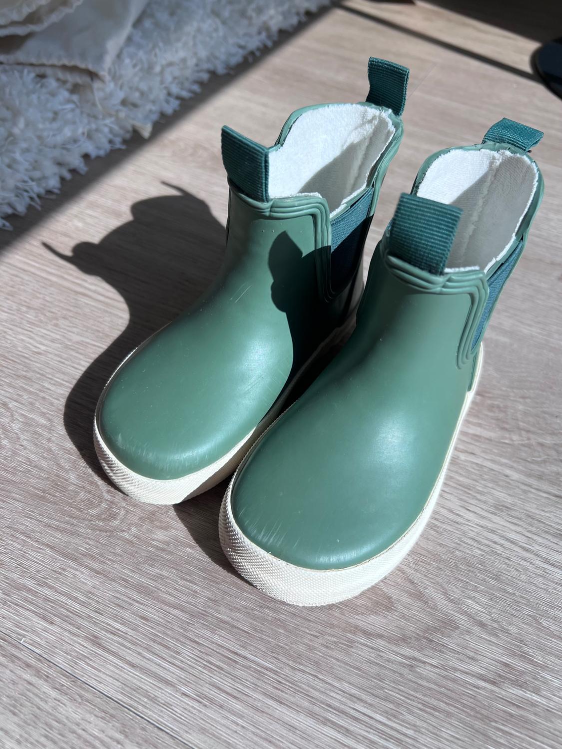 Chelsea-style boots in green with a white rubber sole and an interior lining, placed on a wooden surface under sunlight.