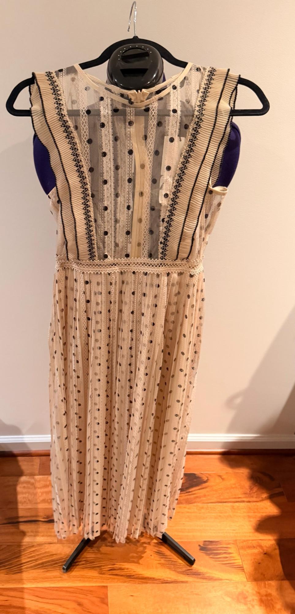Beige dress with black polka dots, lace and pleats, hanging on a mannequin in front of a white wall on a wooden floor.