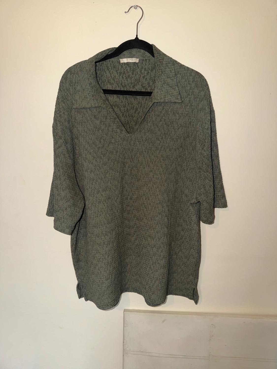 Grayish-green casual short-sleeve polo shirt with a V-neck and textured fabric, hanging on a black hanger against a light-colored wall.