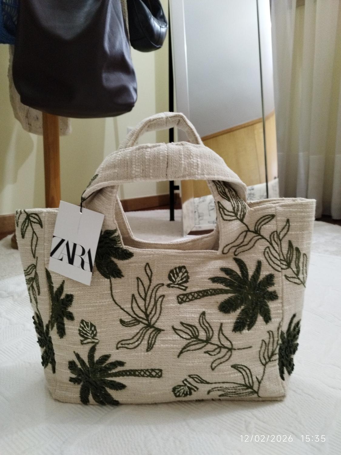 Beige handbag with green leaf embroidery and "ZARA" tag. Background with mirror, curtains and other handbags hanging. Date: 12/02/2026 15:35.