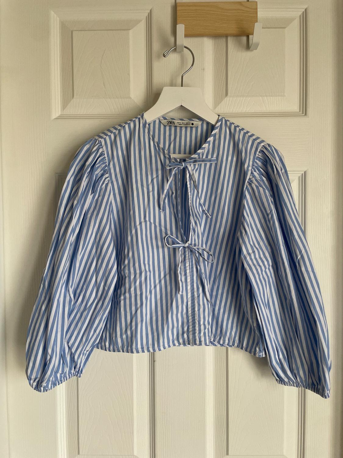 Blue and white striped blouse with a bow at the neck, puff sleeves, hanging on a white hanger over a white door.