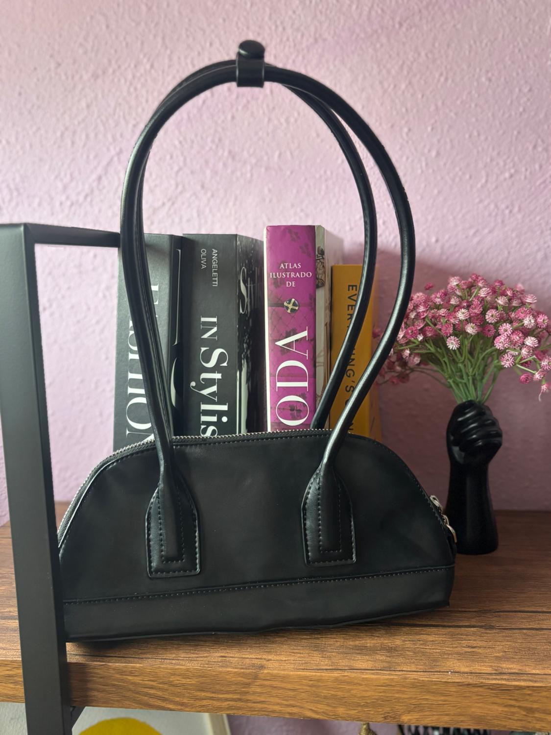 Black handbag with long handles on a wooden shelf, next to books and a black vase with pink flowers, pink wall background.