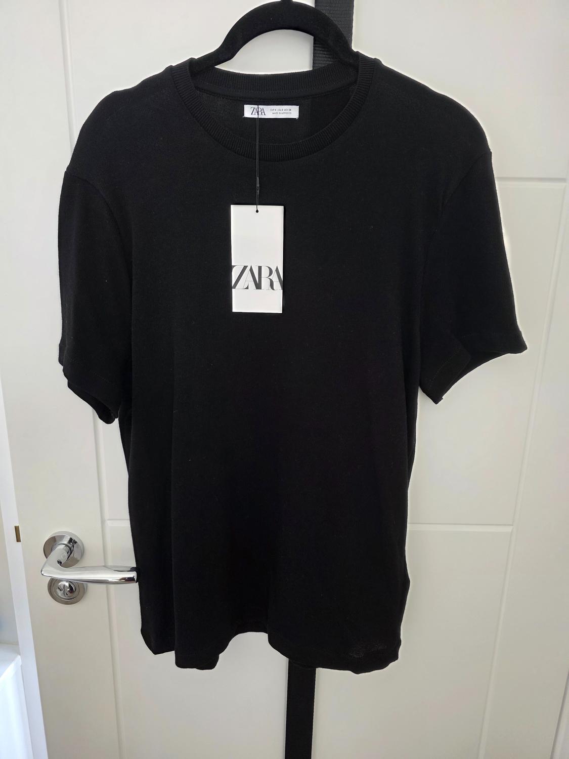 Black T-shirt hanging on a hanger with a white label that says "ZARA", on a white door with a silver handle.