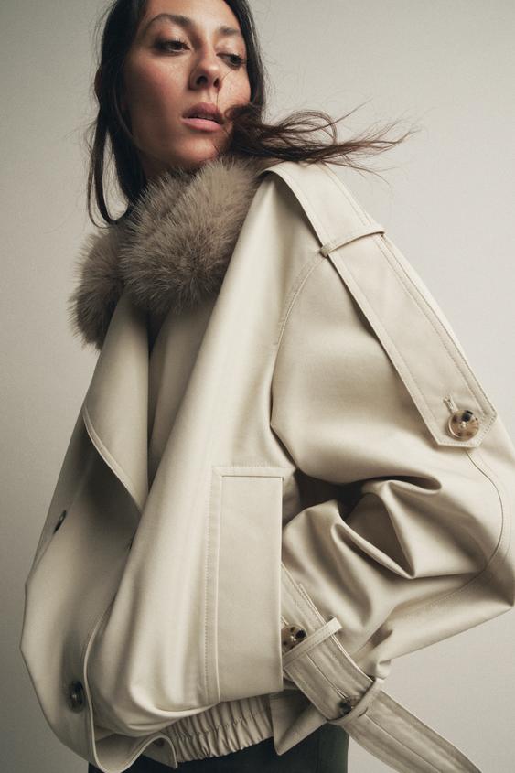 Béžový kabát střední délky s kožešinovým límcem, širokými klopami, přední kapsou a páskem s přezkou.