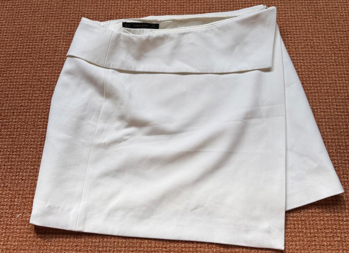 White wrap-over mini skirt with a folded waist on a textured orange background. Label with ZARA BASIC text. Casual flat lay composition.