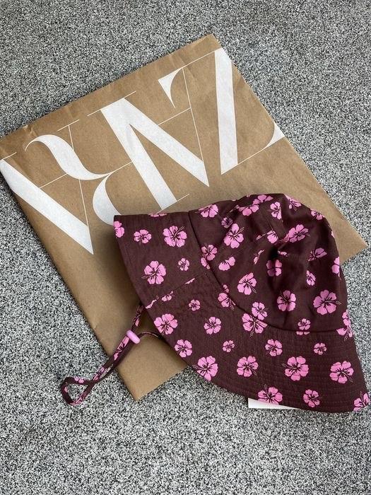 Brown bucket hat with pink flowers and a cord, resting on a paper bag with the text ZARA in white letters on a grey background.