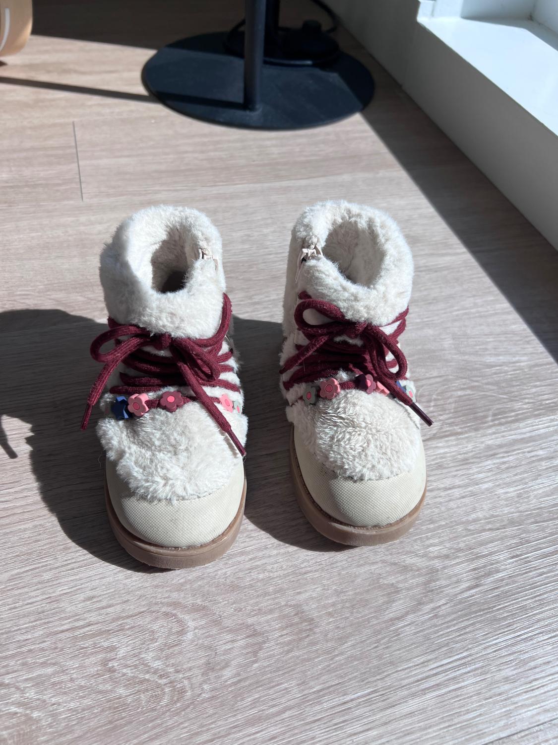 A pair of cream-colored plush-textured ankle boots, burgundy laces and small flower embellishments, on a wooden floor under the sunlight.