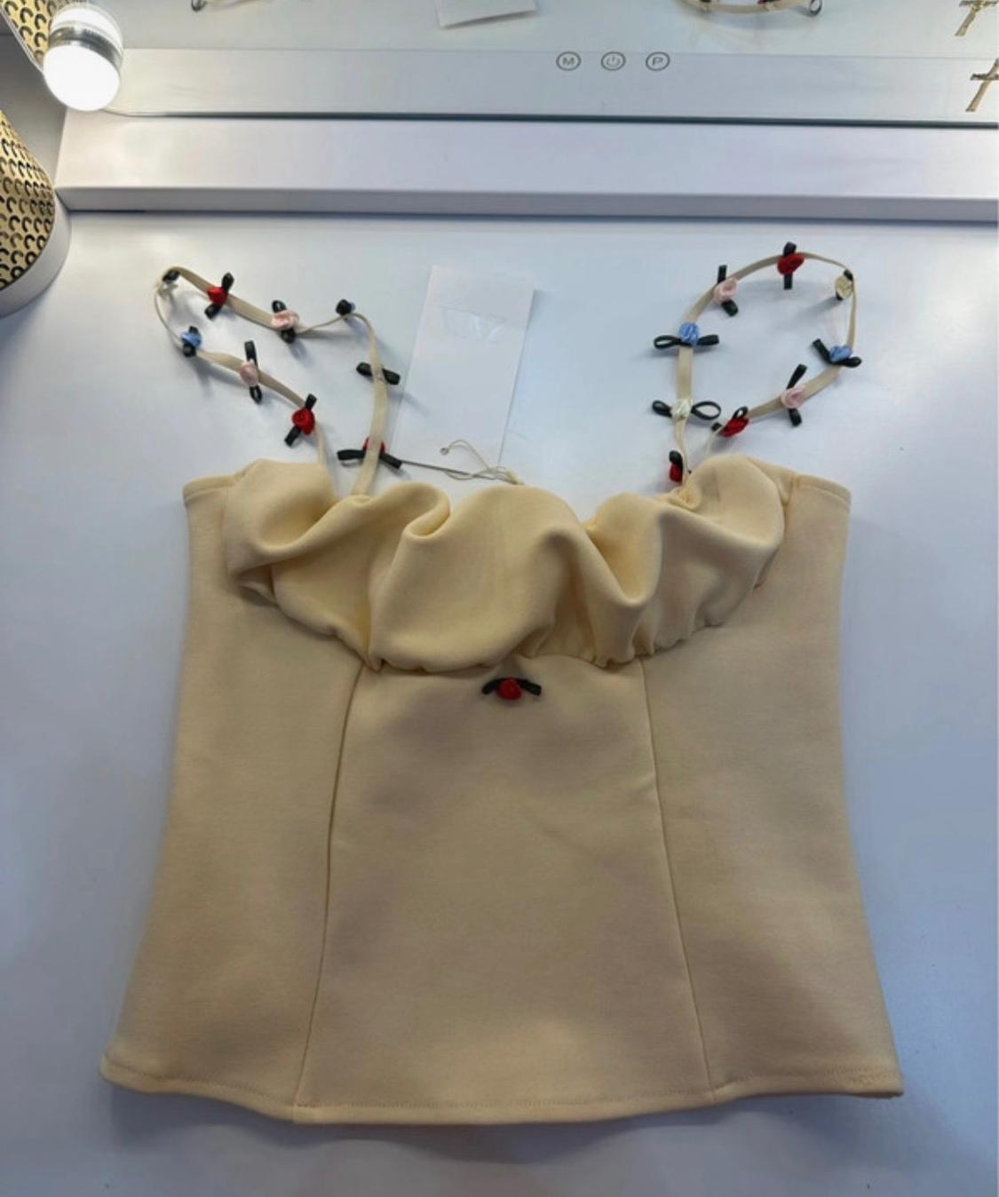 Pale yellow top with a gathered neckline and thin straps decorated with small colourful appliqués, set against a white surface.