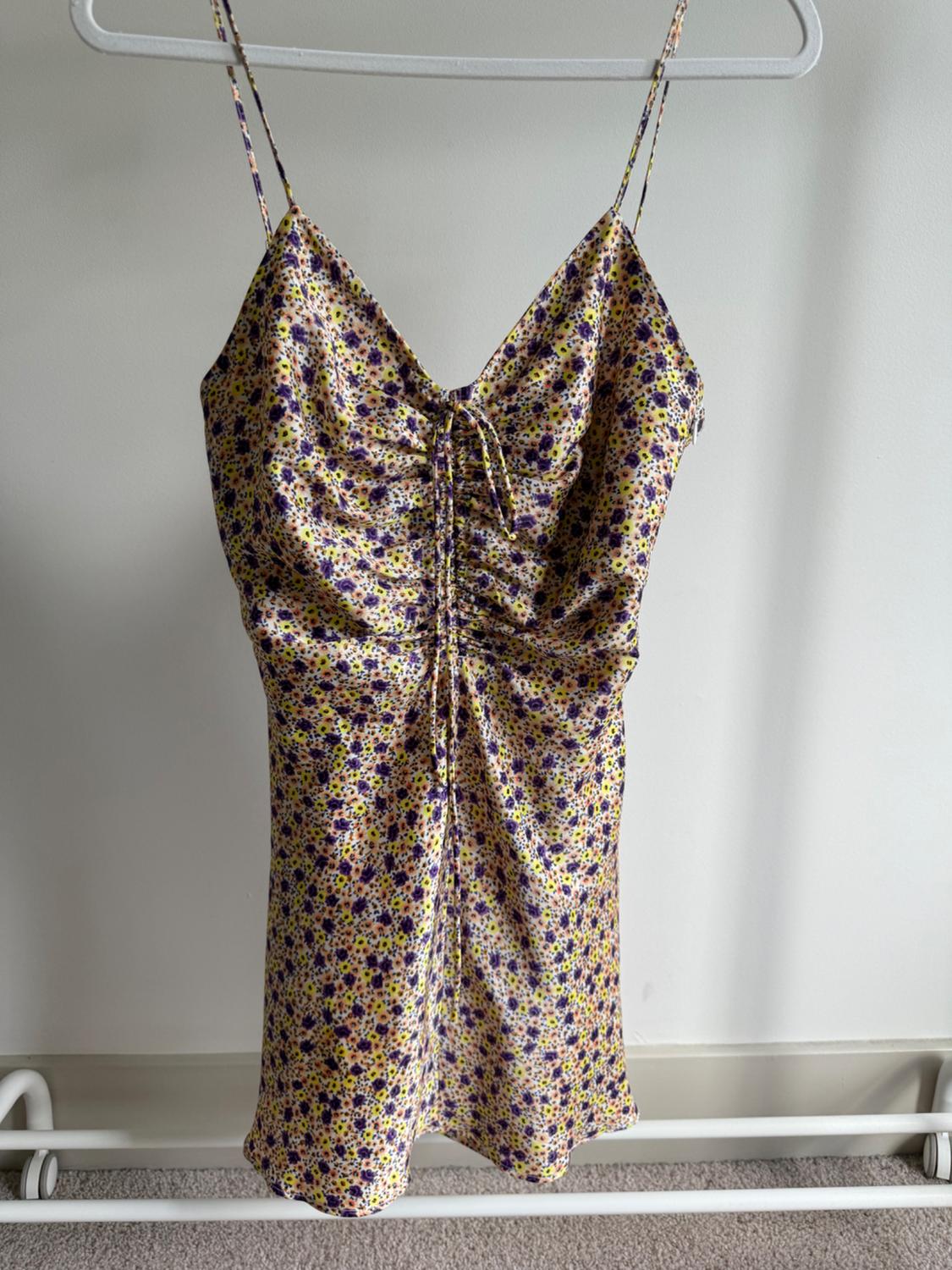 Short sundress with thin straps and a floral print in shades of purple, yellow and white, hanging on a white hanger against a neutral background.