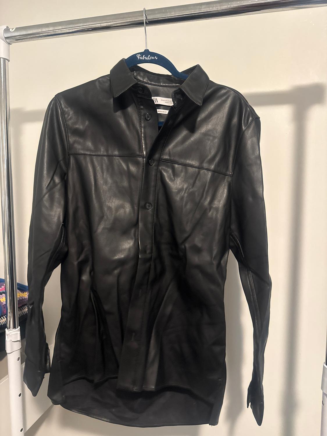 Black leather shirt hanging on a blue hanger with the word "Fabulous", against a white background and a metal clothes rail.