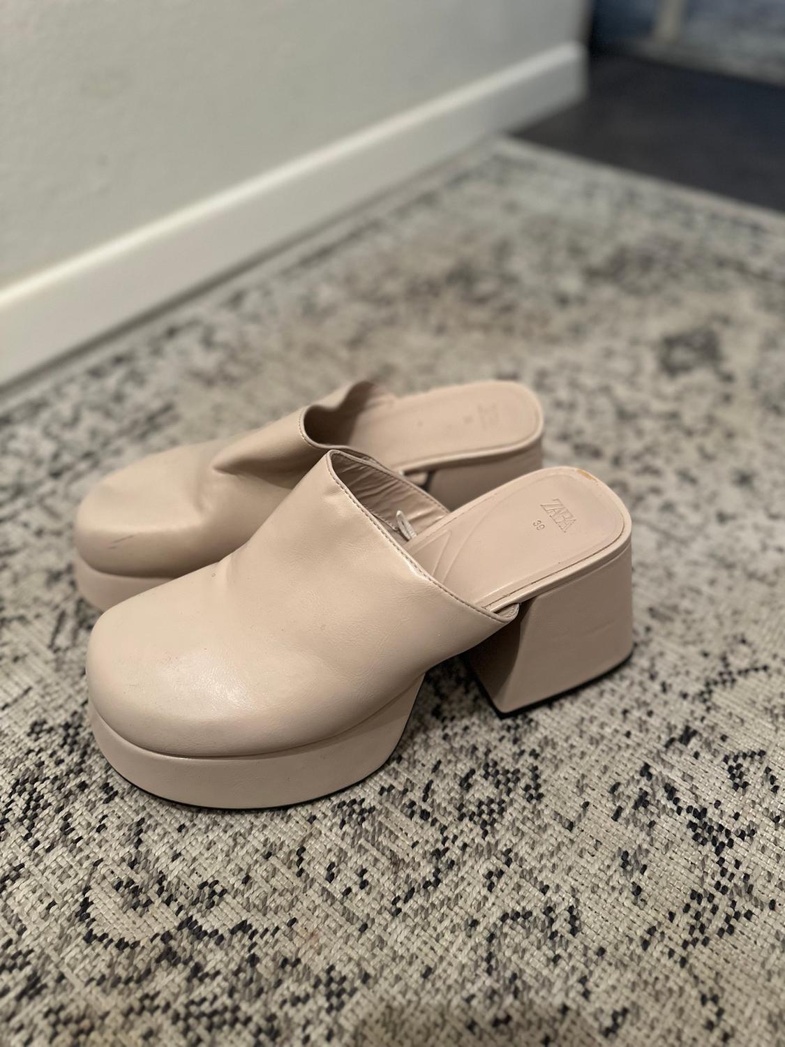 Beige block heels on a patterned grey rug. White wall and dark floor background.
