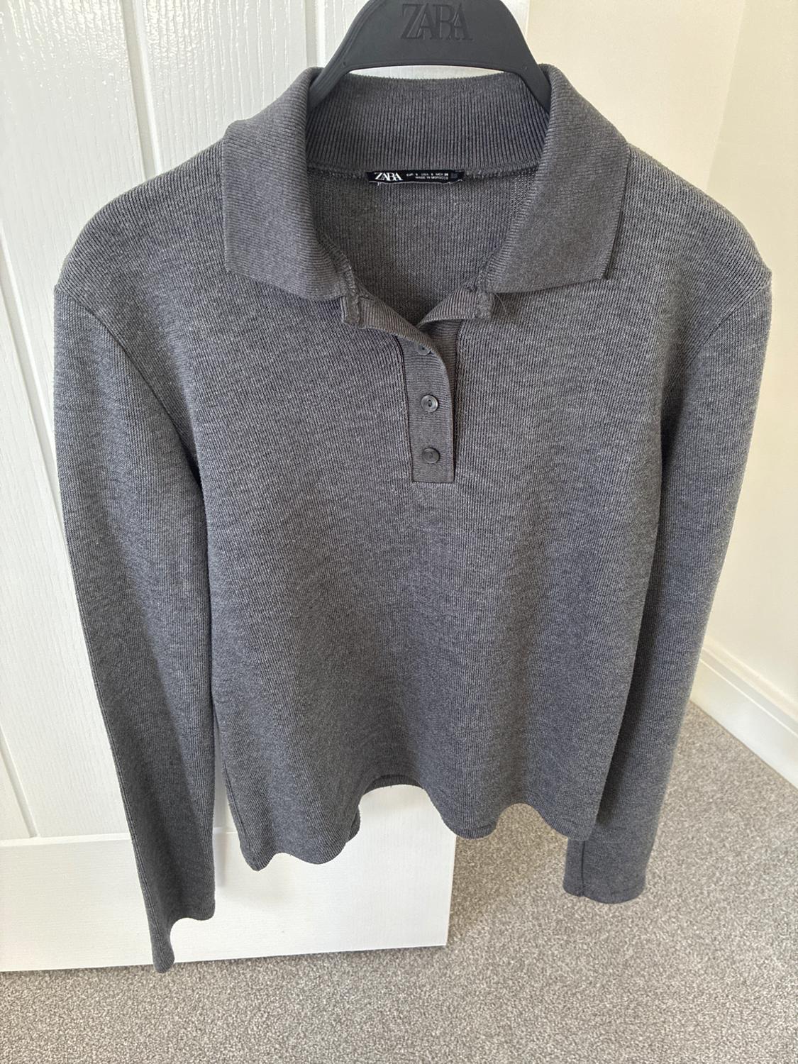 Grey knit polo shirt with long sleeves, a collar and buttons, hanging on a black coat hanger with the text ZARA in front of a white door.