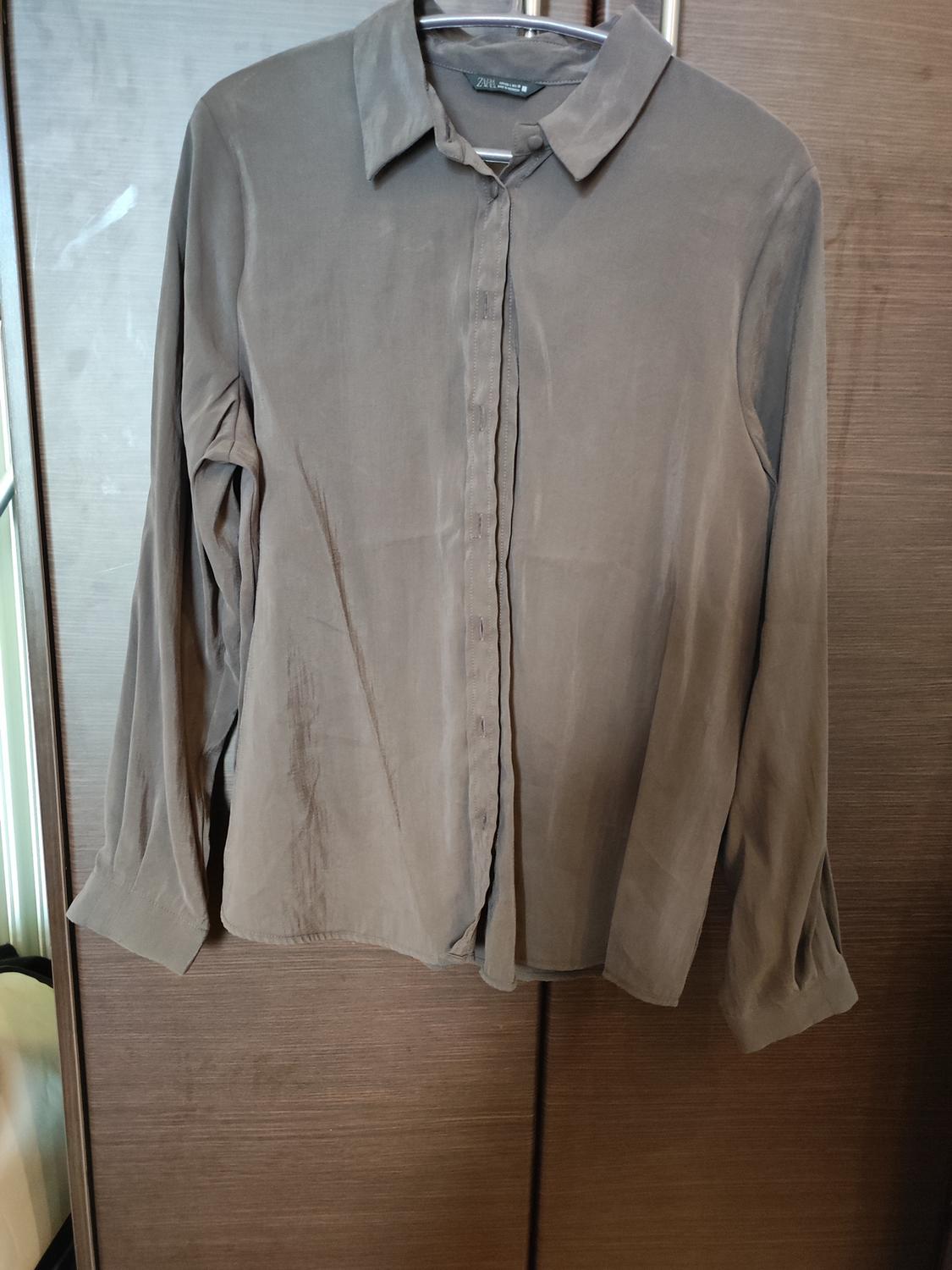 Brown long-sleeved shirt hanging on a hanger, with a wooden background. Label visible on the collar.